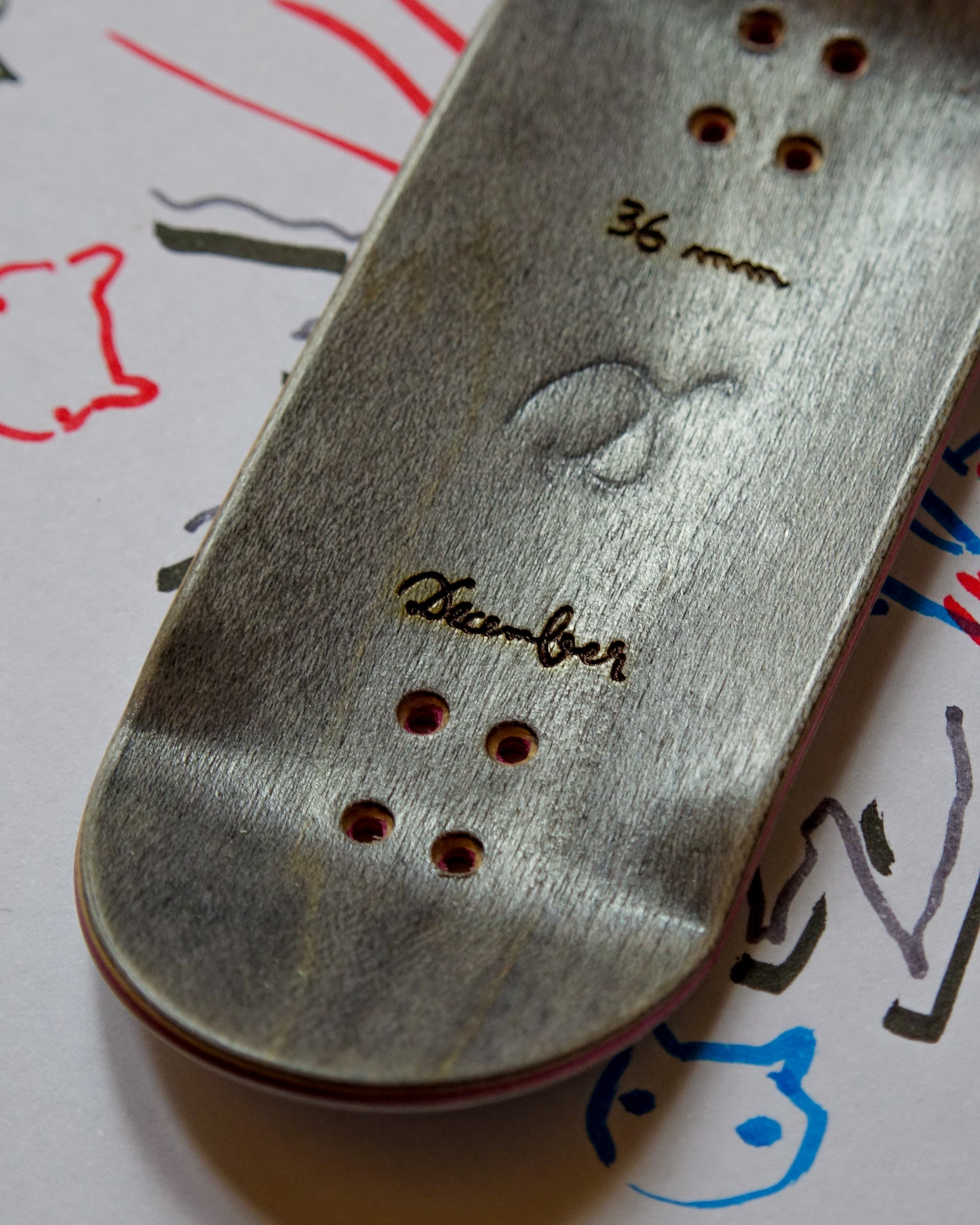 Close-up of a fingerboard deck with December branding on a surface with drawings.